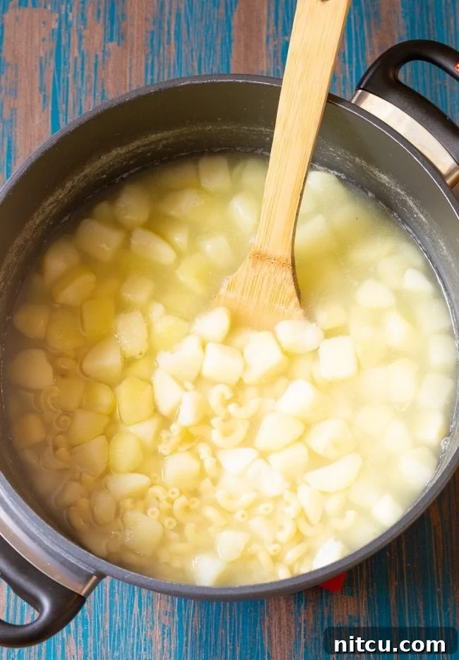 Creamy Hawaiian Macaroni Potato Salad 3 Pasta and potatoes boiling in a pot with a wooden spoon, indicating the start of the cooking process.