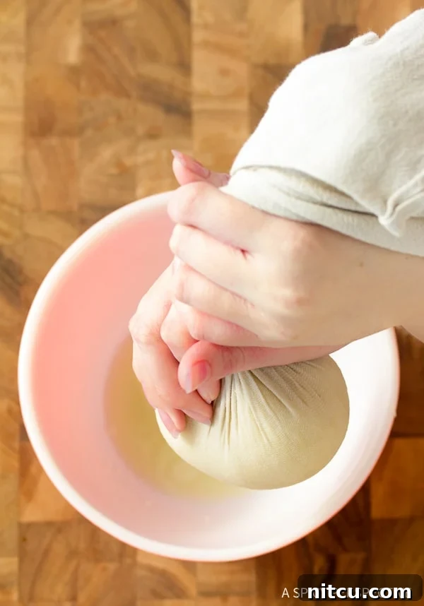 Crispy Keto Cauliflower Bites 6 Hands firmly squeezing excess water from a bundle of steamed cauliflower wrapped in a cheesecloth over a bowl, illustrating a crucial step for crispy crackers.
