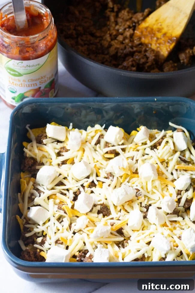 Layers of Doritos and minced meat topped with shredded cheese and cream cheese cubes in a baking dish.