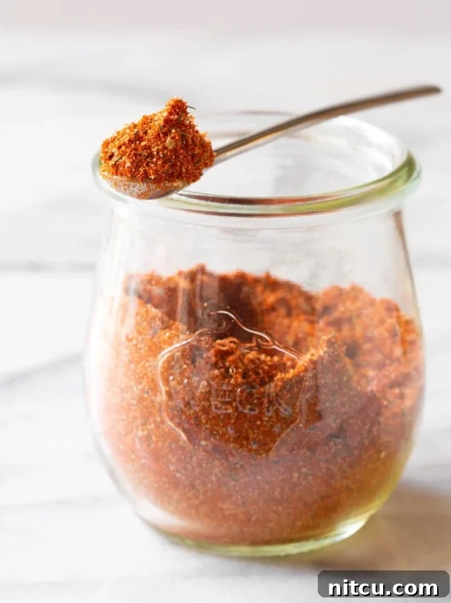 A glass jar brimming with homemade blackened seasoning, with a metal spoon holding a scoop of the spice blend. Perfect for enhancing your cooking.