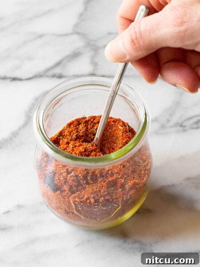 A hand gently holds a metal spoon, poised inside a glass jar filled with our exquisite blackened seasoning, ready to scoop out flavor.