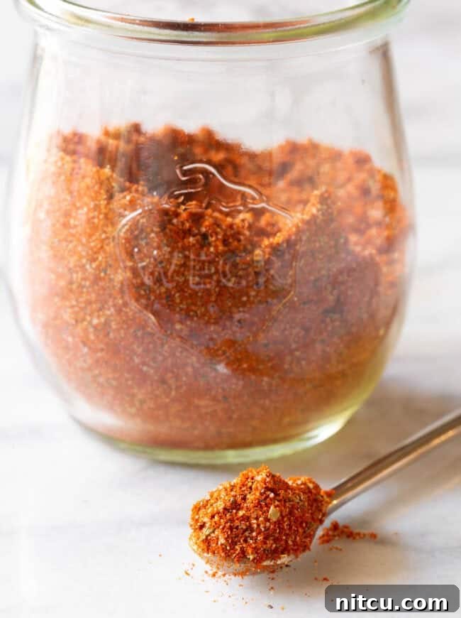 A beautiful glass jar filled with rich, homemade blackened seasoning, with a metal spoon prominently displaying a spoonful of the spice blend in the foreground.