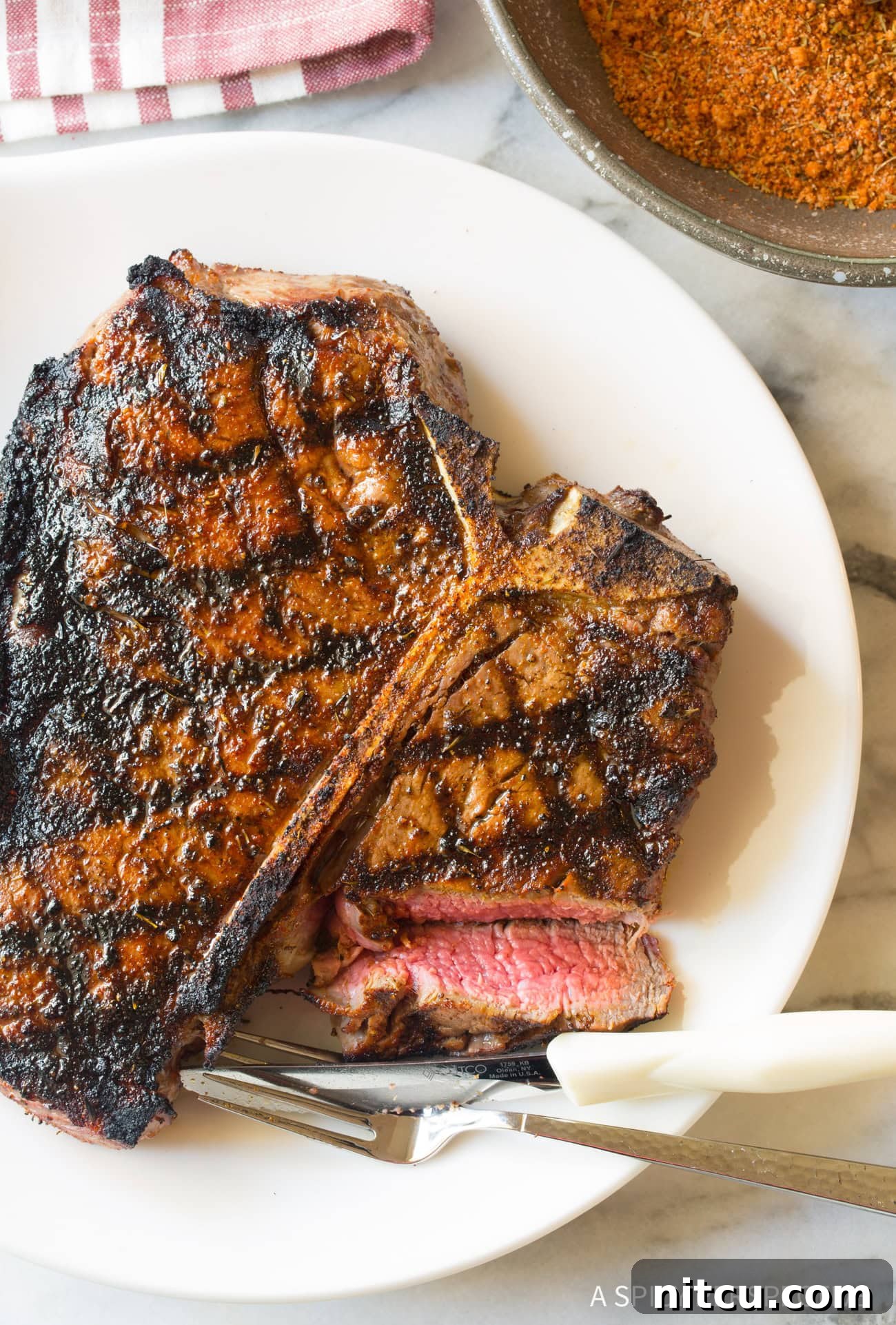 Best Homemade Steak Seasoning Recipe A perfectly cooked steak, glistening with juices, rests on a white plate beside a fork and knife, ready to be enjoyed.