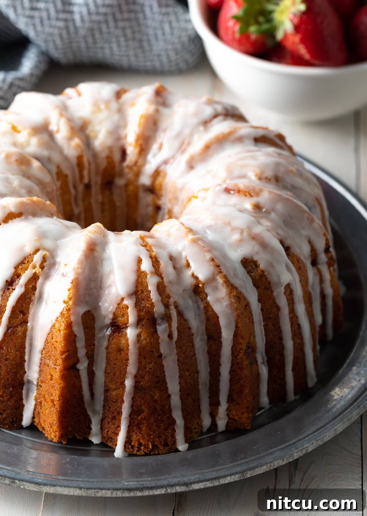 Fresh Strawberry & Lemon Yogurt Bundt Cake A beautifully glazed Strawberry Bundt Cake with lemon drizzle, resting on a elegant silver serving platter. The cake is adorned with fresh strawberry slices, highlighting its vibrant, natural ingredients.