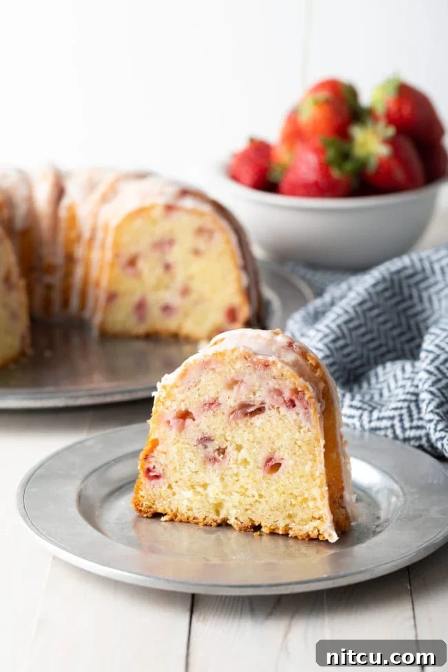 Strawberry Bliss Bundt 13 A close-up of a slice of strawberry bundt cake on a plate with the rest of the cake, beautifully glazed, in the background. The focus is on the inviting texture of the cake.