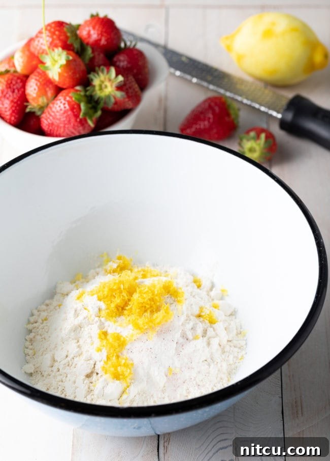 Strawberry Bliss Bundt 5 A mixing bowl filled with sifted flour and lemon zest, with a separate bowl of fresh strawberries beside it on a kitchen counter, preparing for the cake batter.