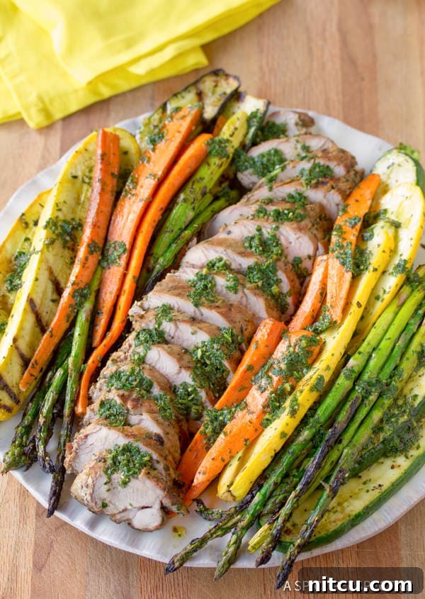 Flame-Kissed Pork Tenderloin with Bright Chimichurri and Rustic Roast Vegetables 7 A large serving tray filled with perfectly sliced grilled pork tenderloin, vibrant green chimichurri sauce in a small bowl, and an abundant assortment of roasted vegetables.