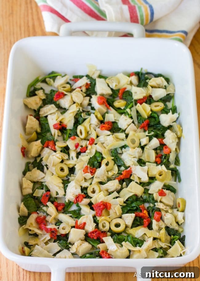 Savory Greek Omelette Bake 5 A casserole dish with vibrant green spinach and tangy artichoke salad evenly layered, awaiting the egg mixture.