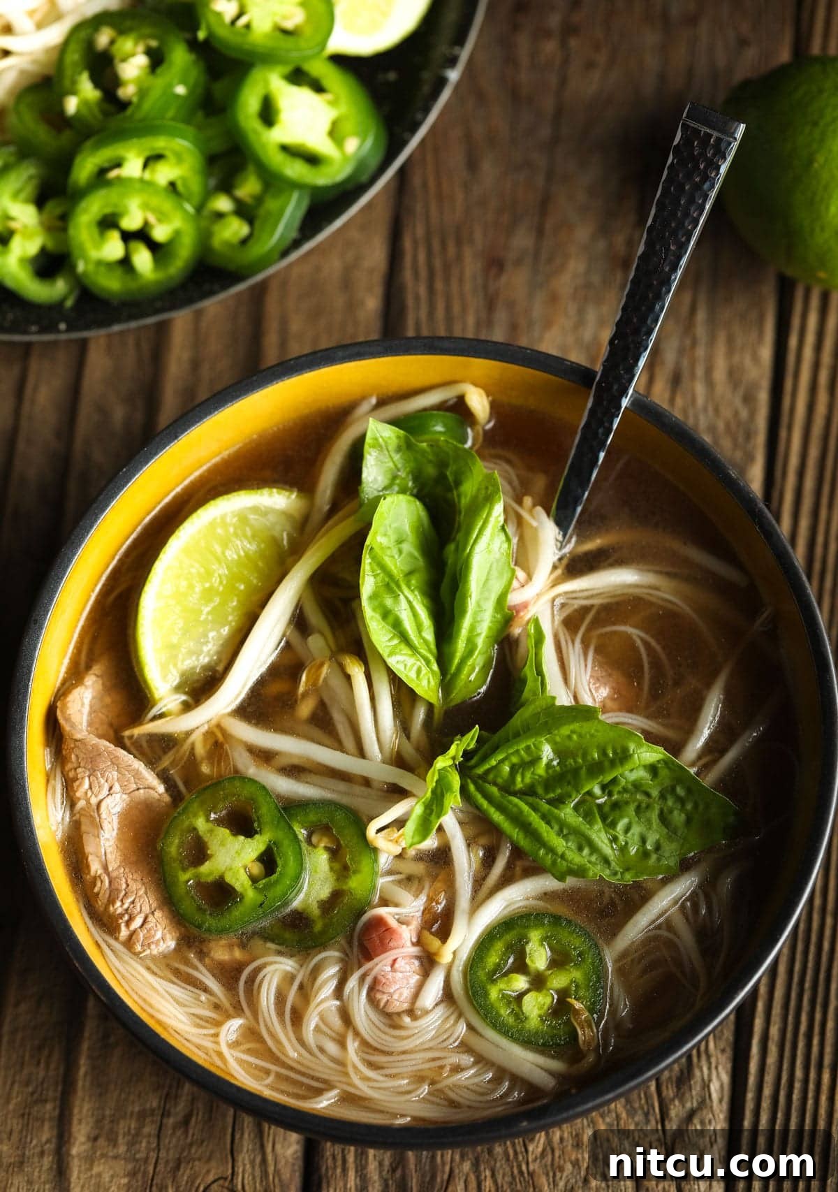 pho-stew Overhead shot of a beautifully arranged healthy pho bowl, adorned with fresh basil, vibrant jalapeños, and a squeeze of lime, showcasing its rich flavors.