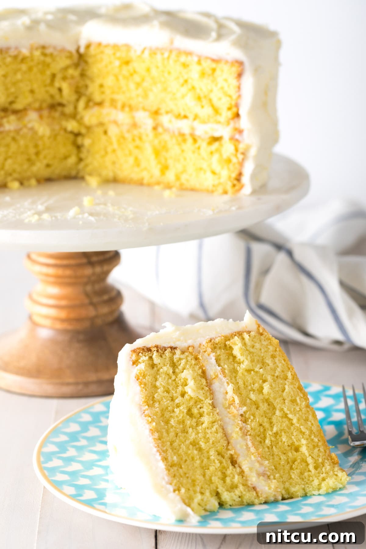 A beautifully sliced lemon buttermilk cake on a blue plate, with the full cake proudly displayed on a stand in the background, ready for serving.