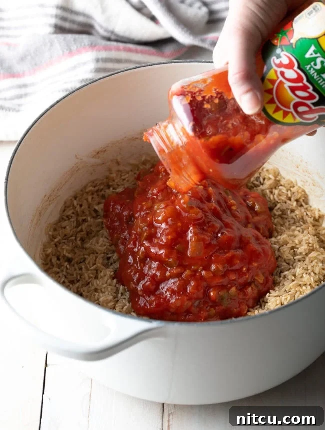 Effortless Spanish Rice 6 Salsa being poured from a jar into a pot with sautéed rice and butter, showing the initial mixing step for the Spanish rice recipe. The vibrant red of the salsa contrasts with the golden rice grains.