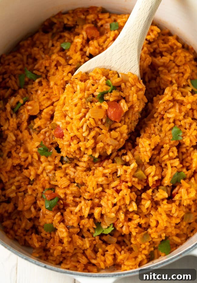 Effortless Spanish Rice 8 A close-up view of a pot of cooked Mexican rice, with a wooden spoon partially submerged, showcasing the texture and vibrant color of the fluffy grains. The steam still rises gently from the rice, indicating it's fresh and warm, ready to be served as a delicious side dish.