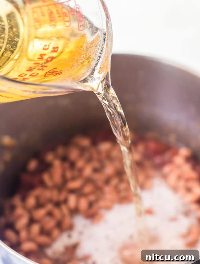 Brewmaster's Chili 5 A measuring cup steadily pours a rich, amber beer into a simmering stock pot filled with chili ingredients, enhancing the depth of flavor.