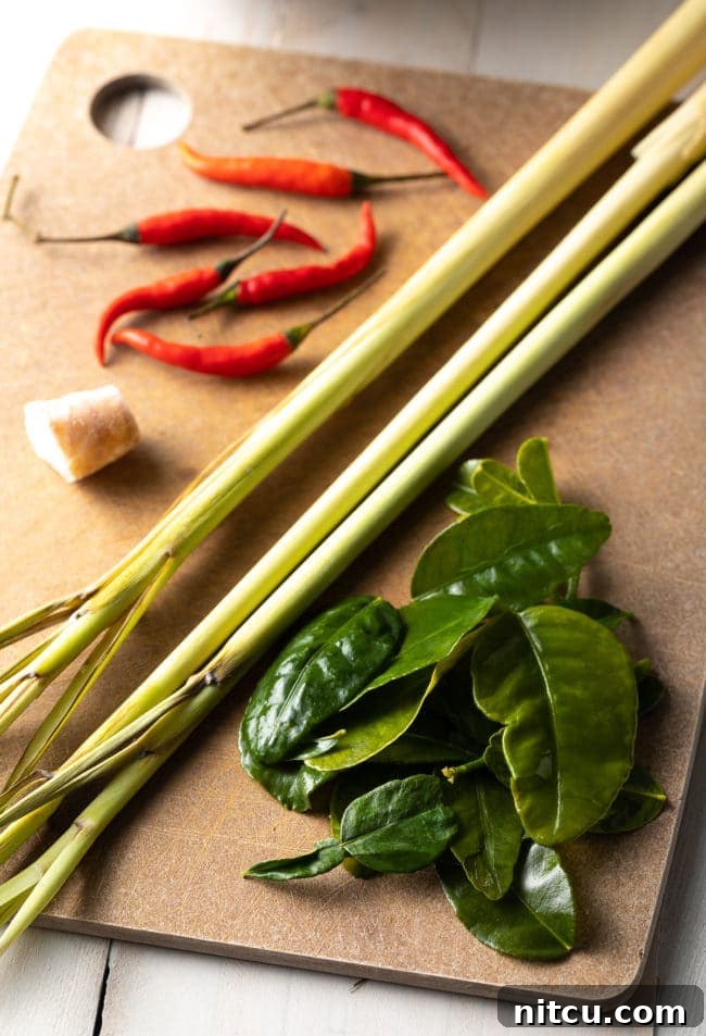 Spicy Thai Tom Yum Soup 6 Fresh ingredients for Tom Yum soup including lemongrass, kaffir lime leaves, galangal, chiles, and onion laid out on a wooden cutting board.