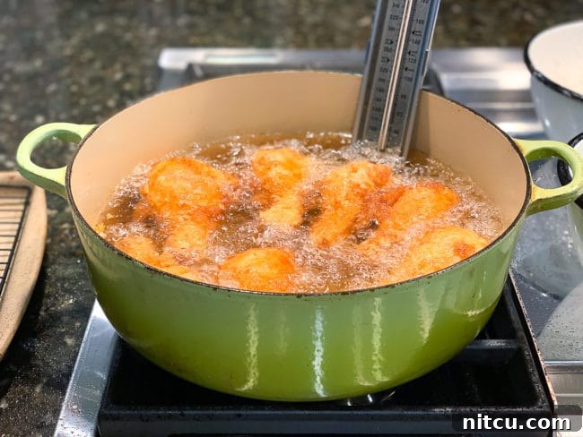 Ultimate Tavern Style Fried Chicken 7 Golden-brown chicken pieces frying in hot oil in a large green Dutch oven, with tongs visible for turning.