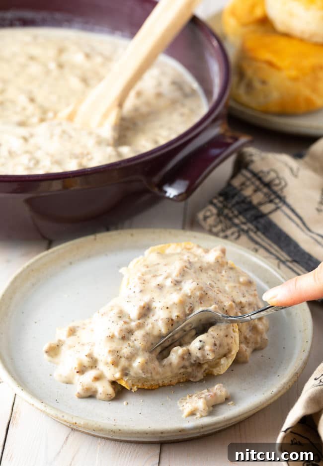 Effortless Homemade Sausage Gravy 4 Hand cutting into a biscuit smothered with spicy sausage gravy on a plate.