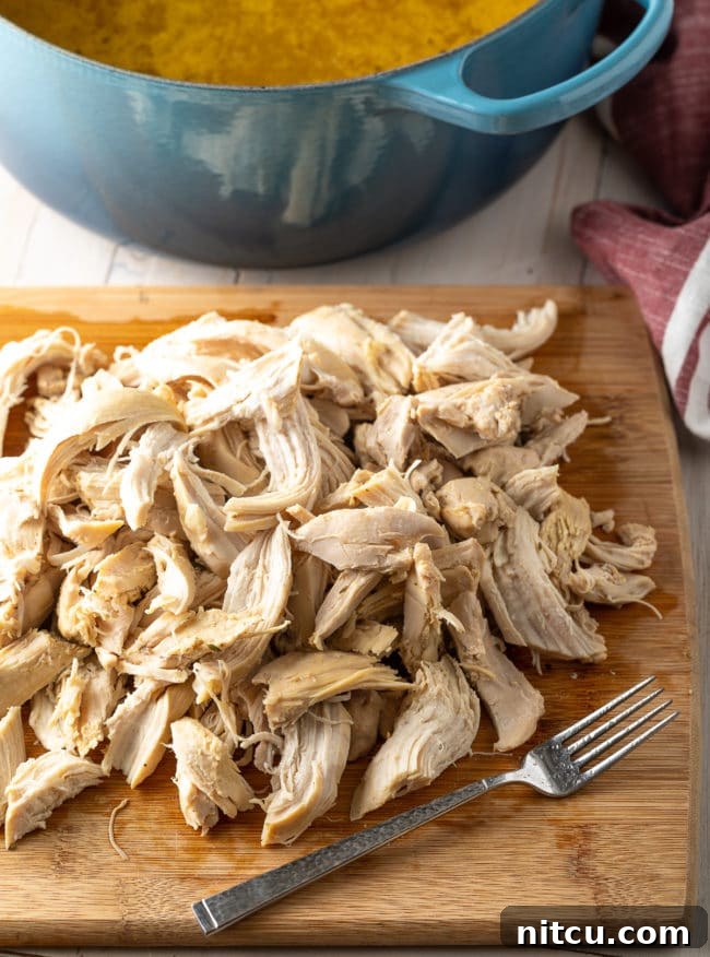 Generous pieces of tender, shredded chicken, freshly pulled off the bone, rest on a wooden cutting board, ready to be added back into the rich chicken and dumpling broth.