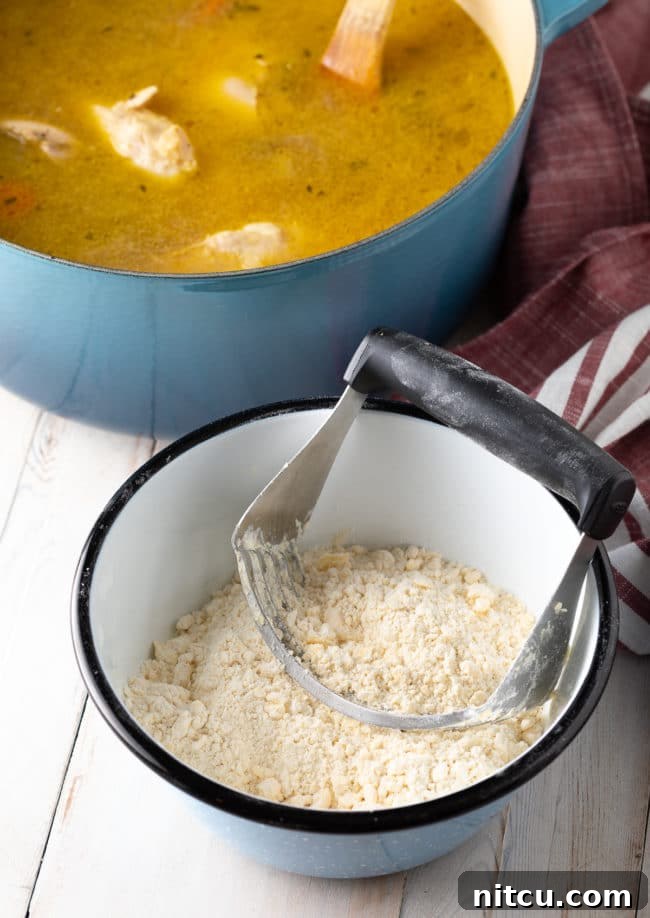 A pastry cutter is used to blend flour and vegetable shortening in a bowl, creating a coarse, crumbly mixture for homemade dumplings, with a blue Dutch oven blurred in the background.