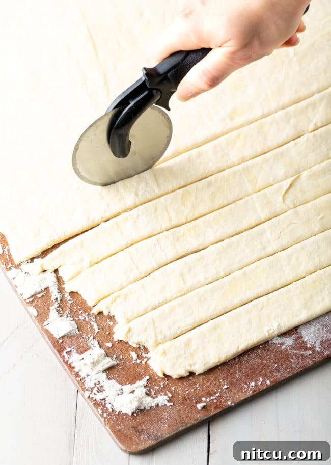 A pizza cutter efficiently slices homemade dumpling dough into long, wide noodle-like strips on a floured cutting board.