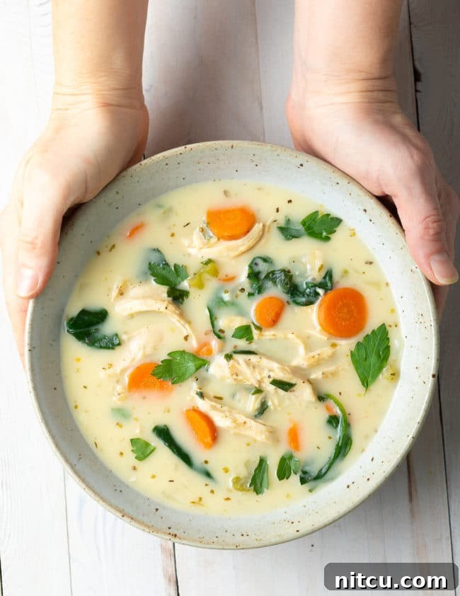Hands holding a speckled bowl filled with creamy lemon chicken soup with vibrant spinach.