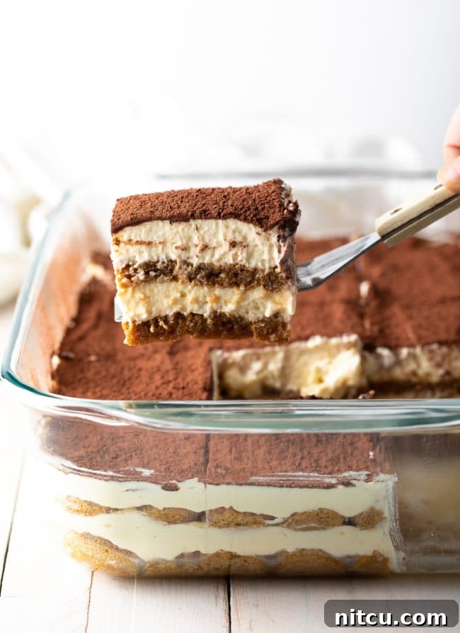 Espresso Pick-Me-Up Delight 2 A perfectly sliced piece of classic Tiramisu held on a spatula, with a larger tray of Tiramisu visible in the background, showcasing its rich layers of cream and coffee-soaked biscuits.