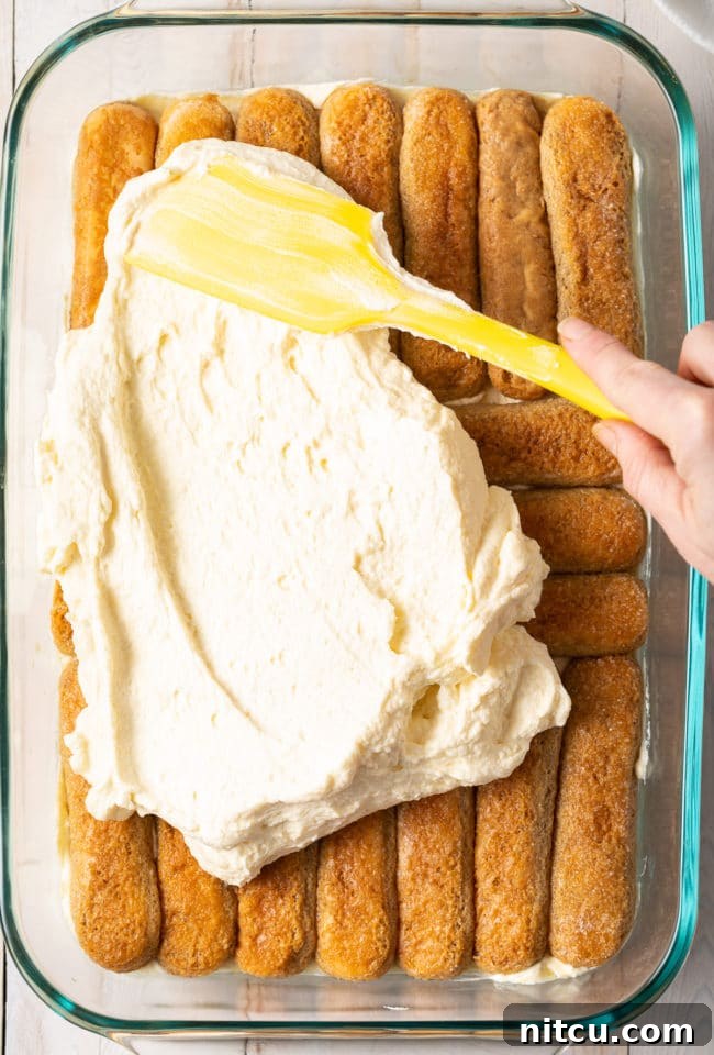 Espresso Pick-Me-Up Delight 10 A spatula smoothly spreading a generous layer of creamy mascarpone mixture over the coffee-soaked ladyfingers in the baking dish, creating the iconic layers.