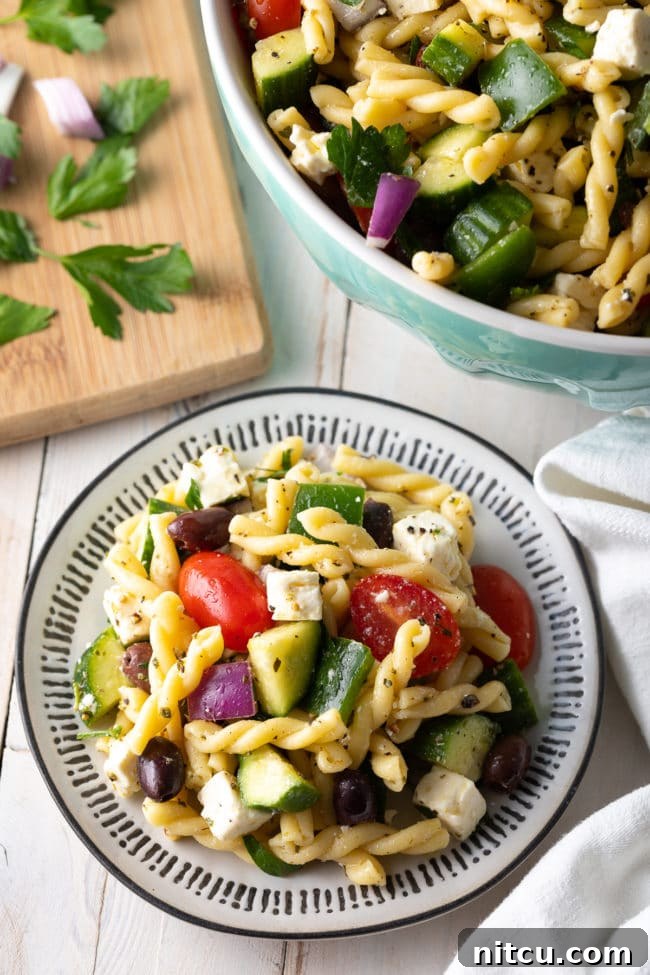 Best pasta salad piled on a white plate, with fresh ingredients like tomatoes, cucumbers, and olives scattered around.