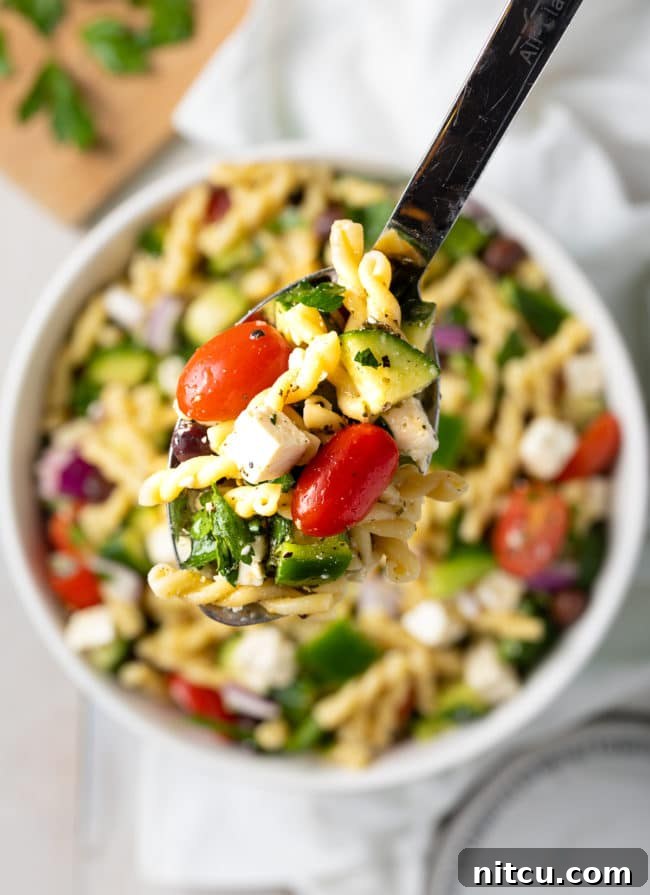 A spoonful of Greek Pasta Salad lifted from a bowl, showing the mix of pasta, olives, tomatoes, and feta.