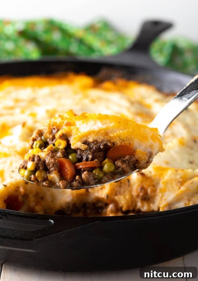 A serving spoon lifting a portion of hot, bubbly Cottage Pie from a cast iron skillet, showcasing the layers.