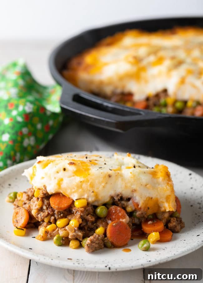 A generous portion of Cottage Pie on a white plate, with a large skillet of the pie blurred in the background, ready to be served.