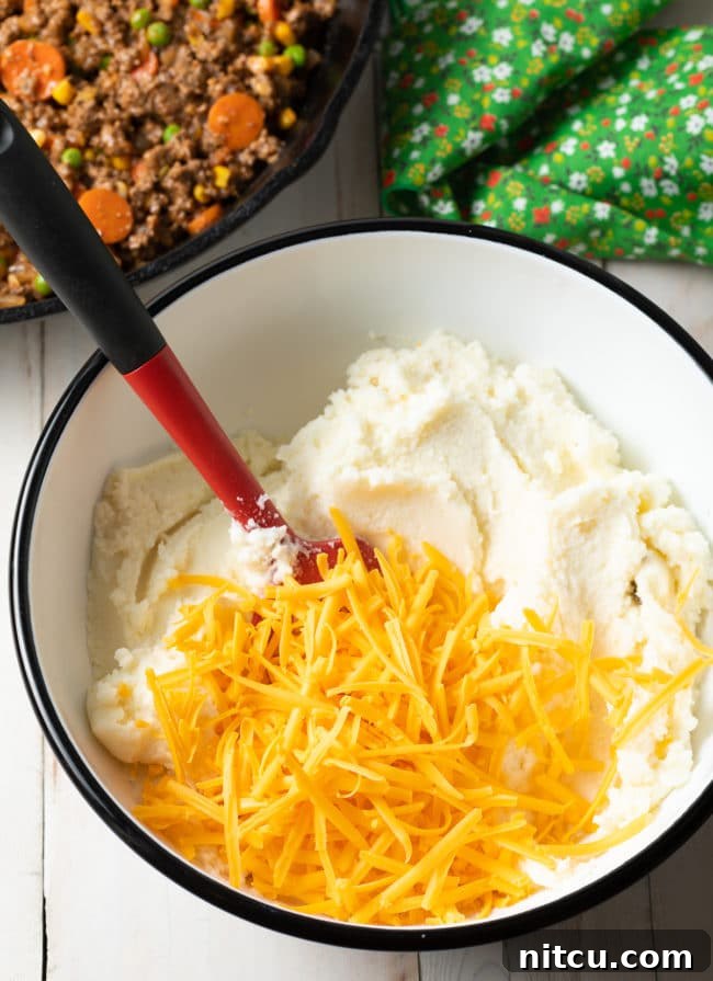 Close-up of creamy mashed potatoes being mixed with shredded cheddar cheese, ready to top the Cottage Pie.