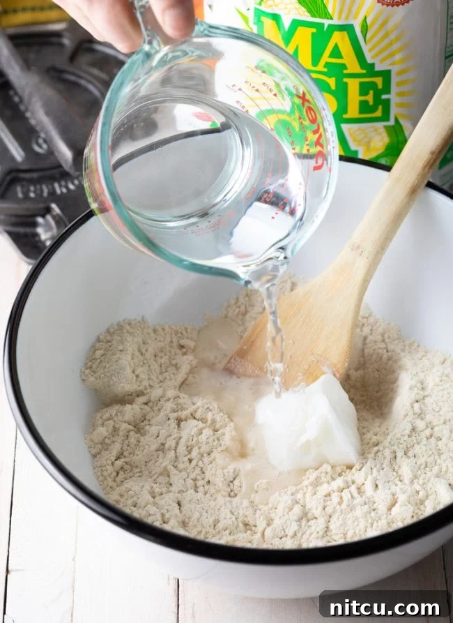 Very hot water being carefully poured into a mixing bowl containing masa harina and lard, initiating the dough-making process.
