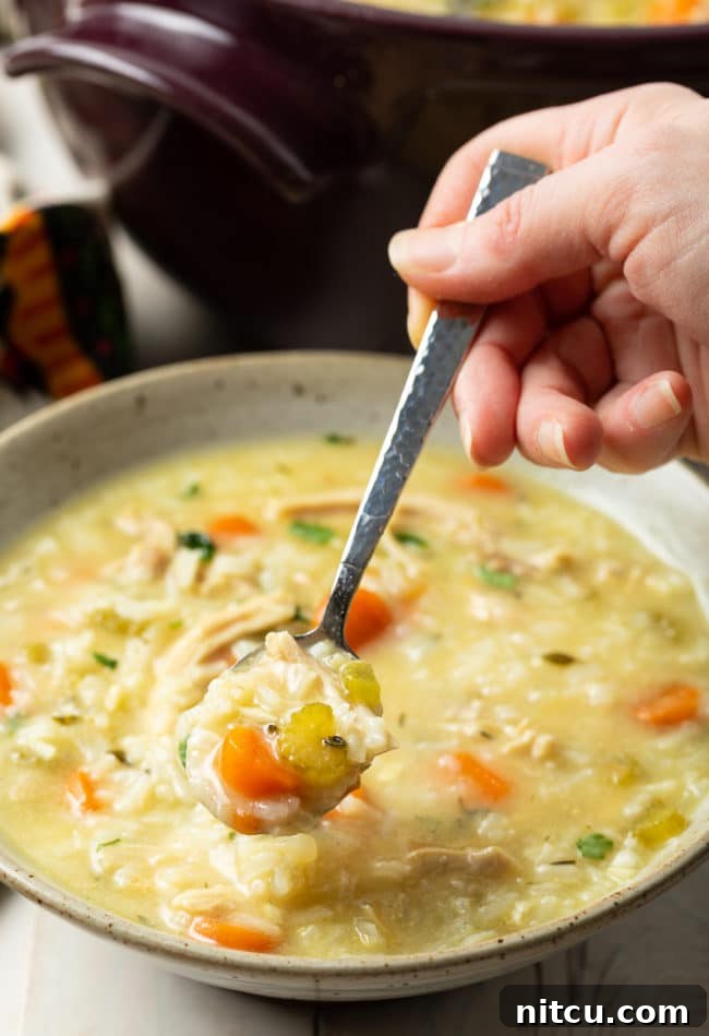 Velvety Chicken and Rice Soup 9 A hand holding a spoon with a spoonful of creamy chicken and rice soup, garnished with fresh herbs, above a steaming bowl.