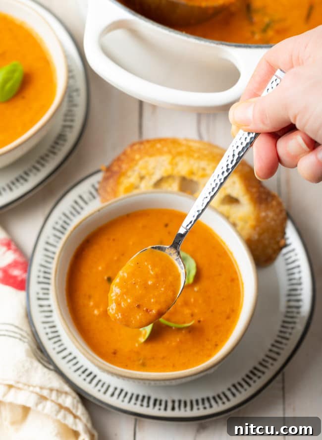Velvety Roasted Tomato and Basil Soup 11 A bowl of creamy roasted tomato soup with a hand holding a spoonful of the soup above the bowl, ready to be tasted.