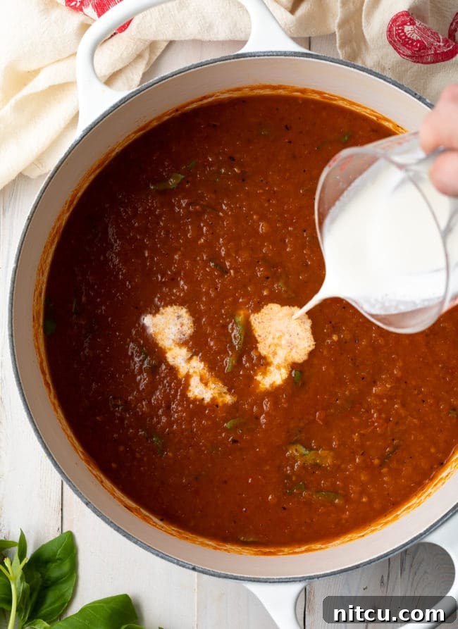 Velvety Roasted Tomato and Basil Soup 9 A hand gently pouring heavy cream into a simmering pot of rich tomato soup, adding to its creamy texture.