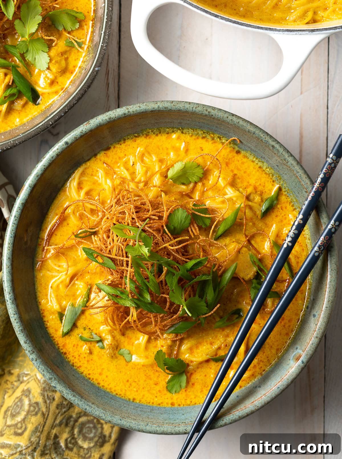 A top-down view of a bowl of rich, golden Khao Soi, garnished with fresh cilantro, sliced red onion, and crispy fried noodles. Chopsticks rest on the bowl's rim, inviting you to dive in.