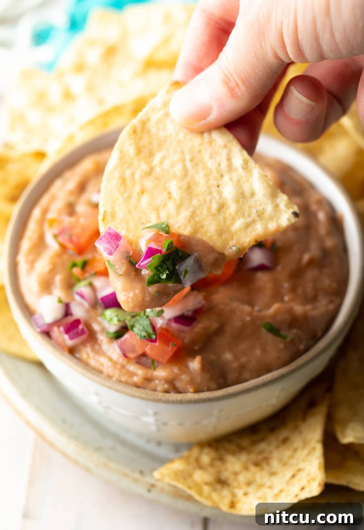 Ultimate Homemade Bean Dip 5 A hand holding a golden tortilla chip, poised to scoop a generous portion of creamy, flavorful homemade bean dip.