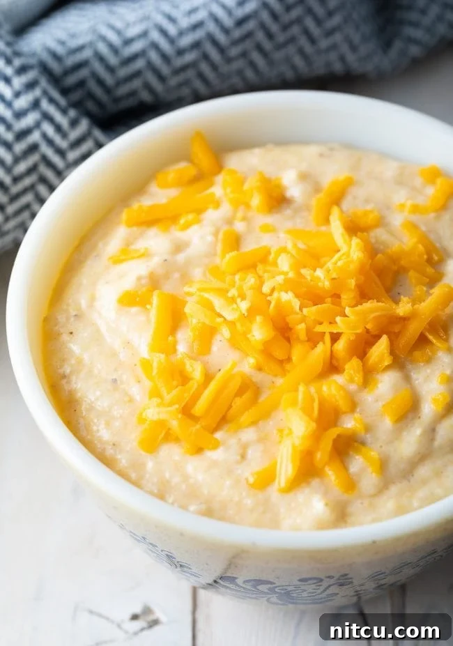 Golden Cheesy Comfort 2 Creamy Cheese grits recipe in a white bowl with shredded cheddar cheese on top.