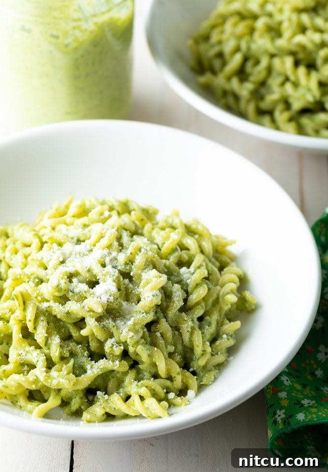 Velvety Pesto Perfection 9 Two white bowls filled with creamy pesto pasta, alongside a jar of the fresh pesto sauce, arranged on a light surface.