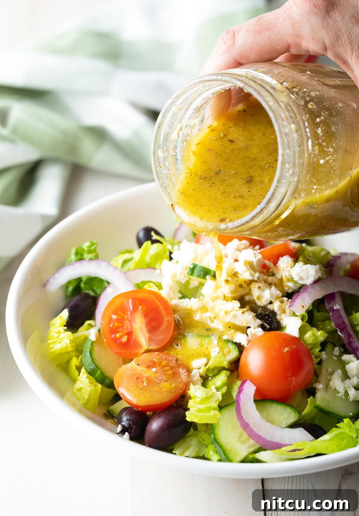 Zesty Greek Vinaigrette 9 A hand pouring a glass of Greek Salad Dressing over a Greek Salad, ready for serving.