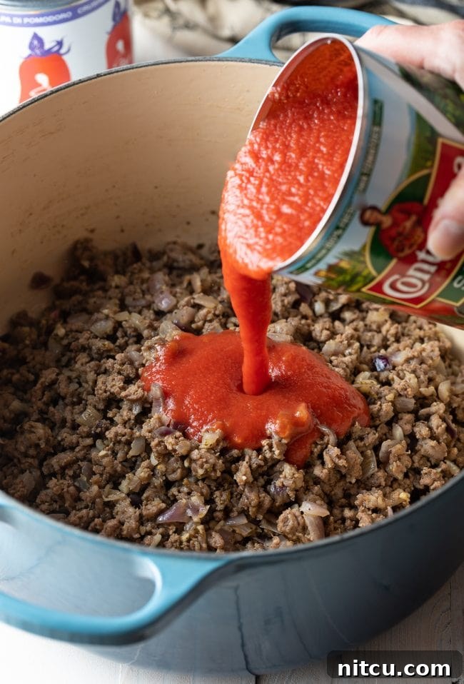 The Ultimate Spaghetti and Meat Sauce 5 Hand pouring a can of tomato sauce into a pot with ground beef.