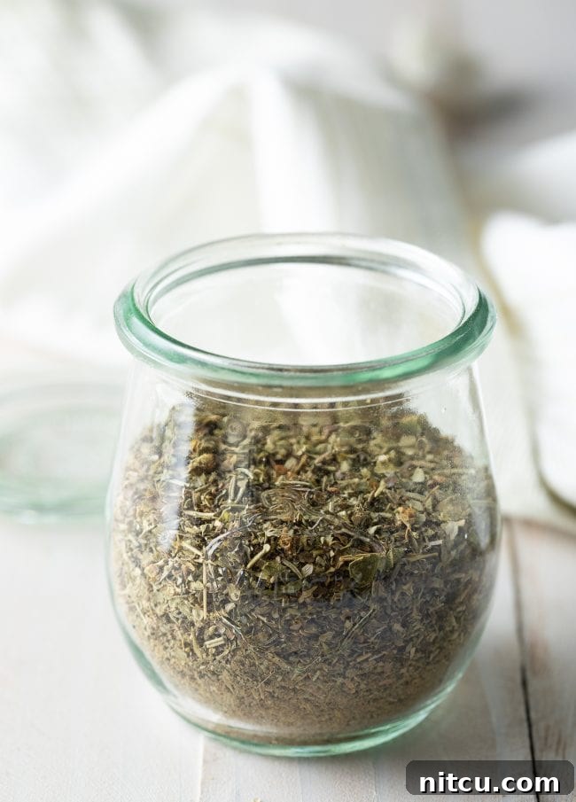 Your Signature Italian Spice Blend 5 A hand holding a spoon filled with homemade Italian seasoning over a jar of the blend.