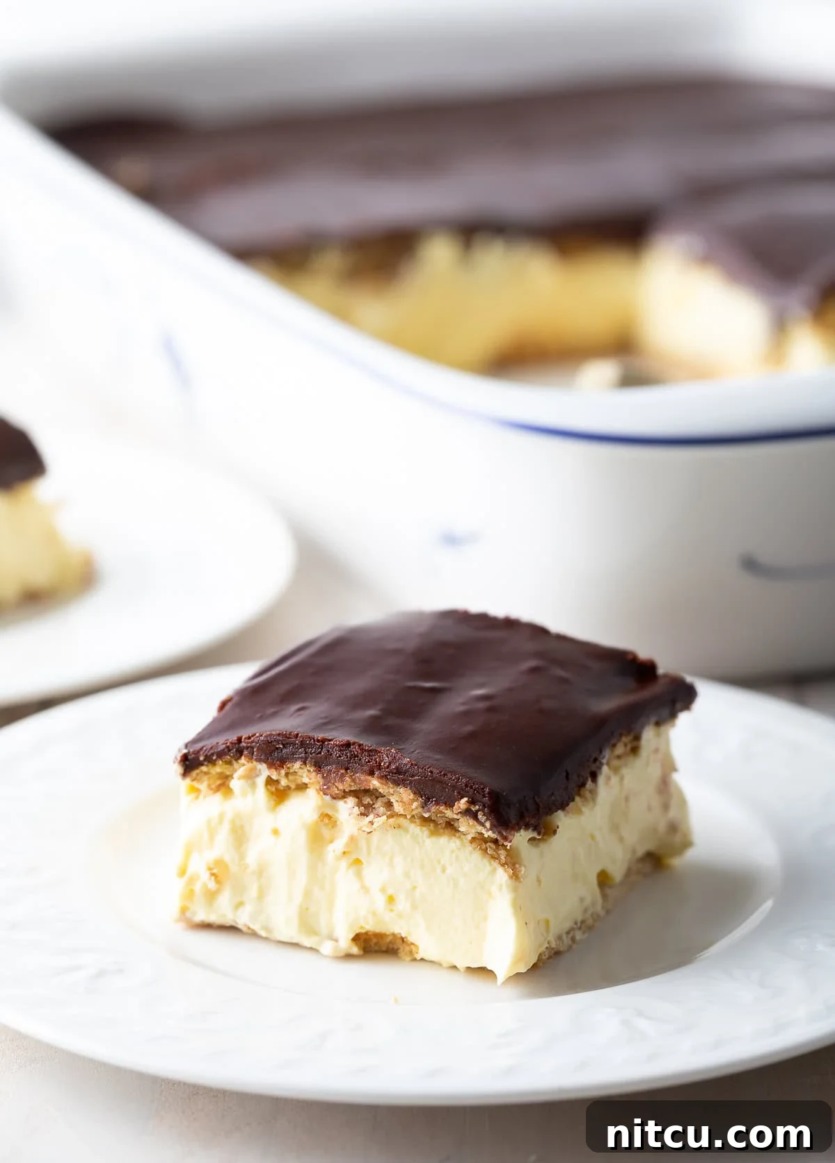 Decadent Chocolate Ganache Eclair Cake 2 A perfectly portioned slice of no-bake eclair cake, showcasing its distinct layers of softened graham cracker, creamy vanilla pudding, and rich chocolate ganache, artfully presented on a white plate, ready to be enjoyed.