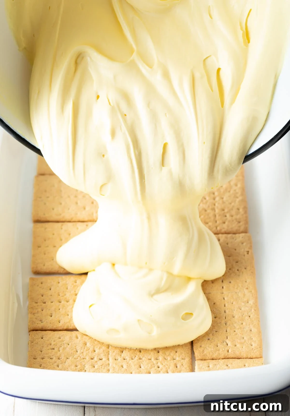 Decadent Chocolate Ganache Eclair Cake 6 A baking tray meticulously lined with a single layer of graham crackers, with the smooth, creamy pudding mixture being poured evenly over them, forming the luxurious second layer of the no-bake eclair cake.