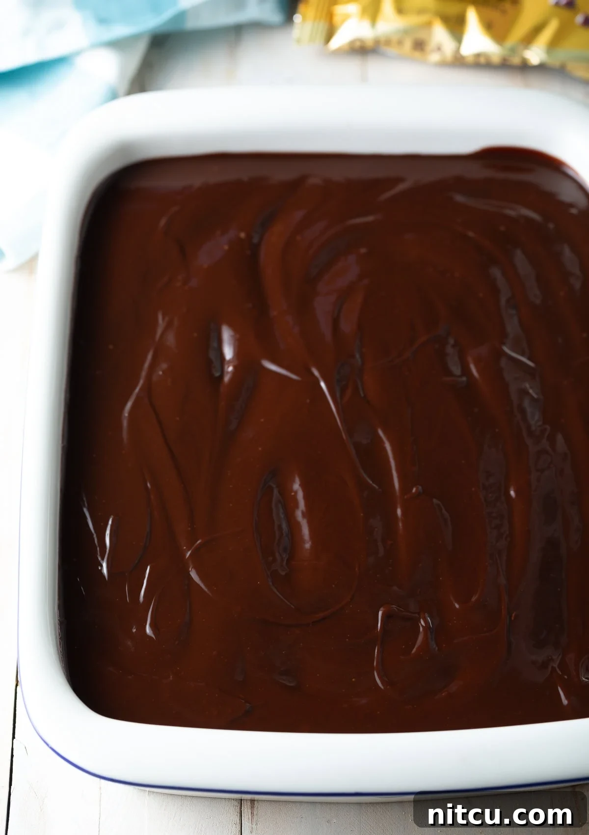 Decadent Chocolate Ganache Eclair Cake 7 A top-down view of the no-bake eclair cake nestled in a baking dish, freshly topped with a rich, glossy chocolate ganache, which is being smoothly spread to cover the entire surface, promising a decadent finish.