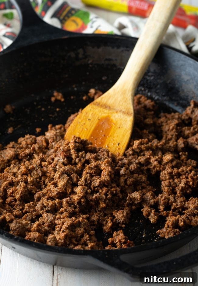 Browning Ground Beef for Macaroni Ground beef browning in a large skillet, being broken apart with a wooden spatula.
