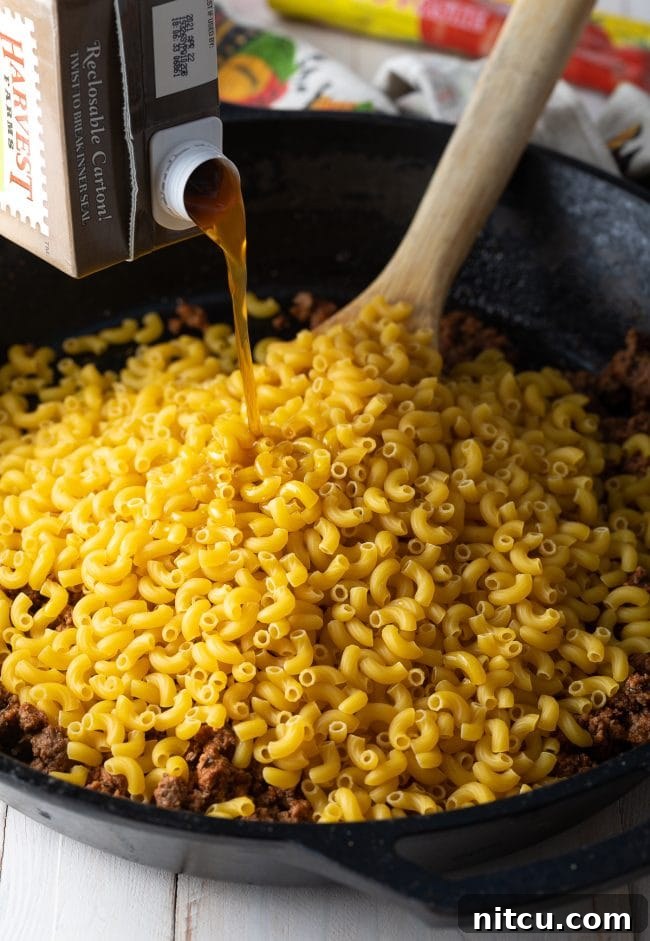 Adding Broth and Macaroni to Skillet Beef broth being poured from a carton into a skillet with uncooked macaroni noodles and seasoned ground beef.