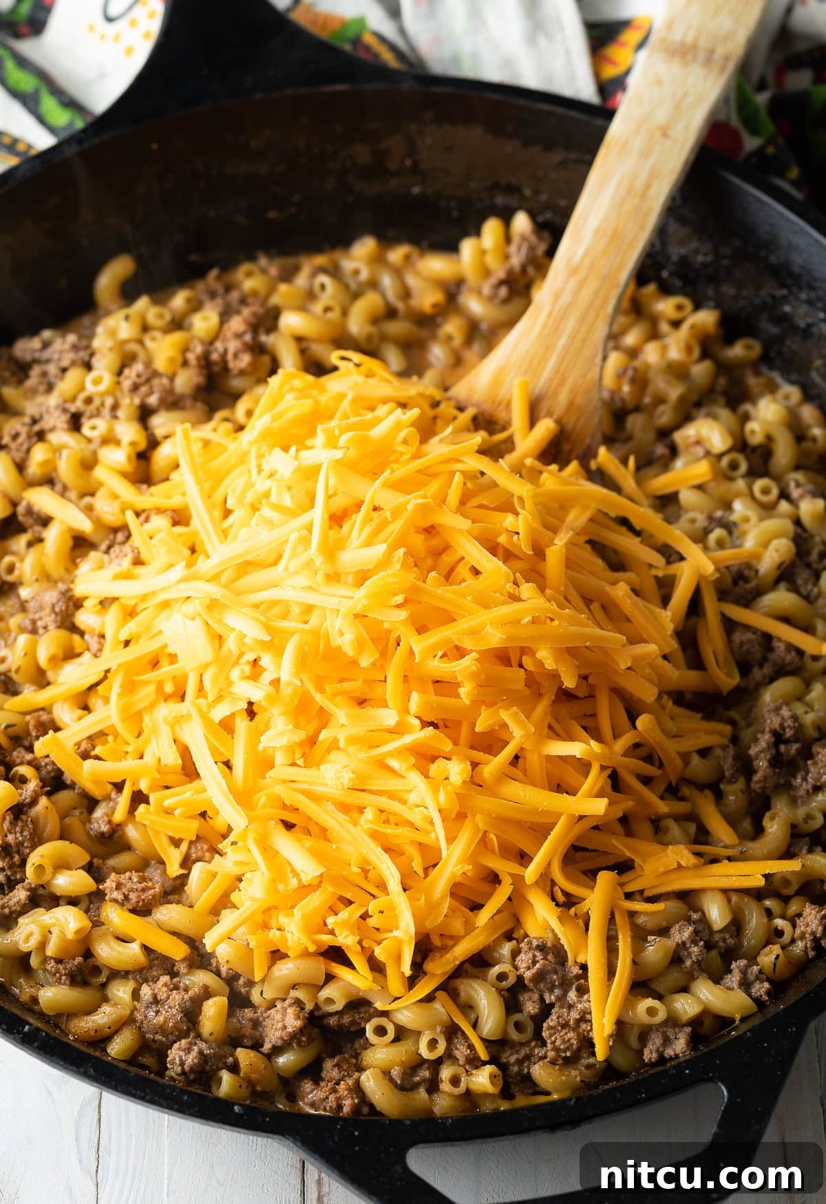 Melting Cheese into Macaroni A hand stirring shredded cheddar cheese into the simmering beef and macaroni skillet, creating a creamy sauce.