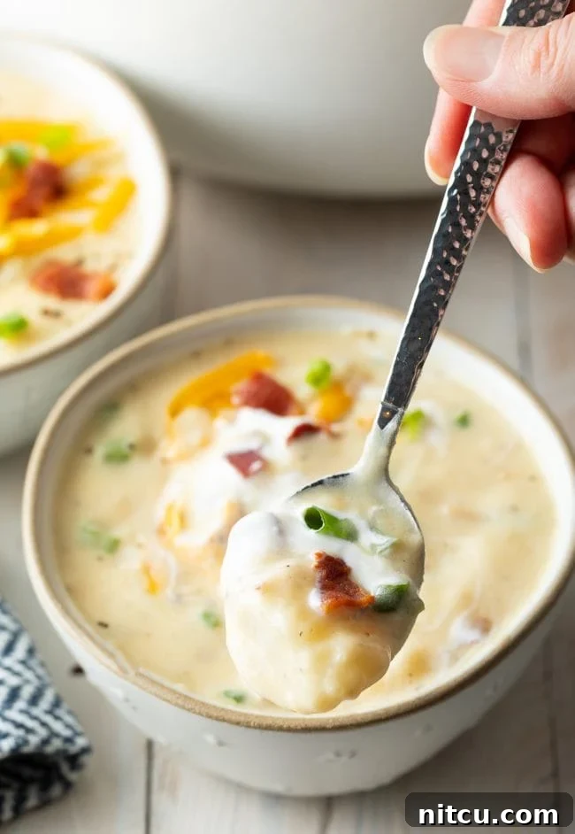 A close-up view of a bowl of chunky potato soup, with a hand holding a spoon poised to scoop up a delicious portion.