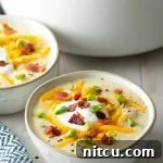 A close-up of a rustic white bowl brimming with hearty loaded baked potato soup, adorned with a swirl of sour cream, golden cheese, and fresh green onions.